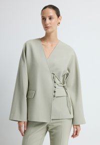 Jasnozielona kurtka typu wrap z szerokimi rękawami, głębokim dekoltem w szpic i ozdobnymi sznurkami po bokach. Wykonana z gładkiego materiału.