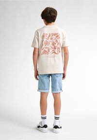 Béžové bavlněné tričko s růžovým květinovým designem na zádech, spárované s světle modrými denimovými šortkami a bílými teniskami s černými akcenty.