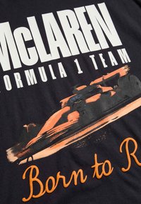 Svart t-shirt med vit text "McLaren Formula 1 Team", stiliserad F1-bil i orange och orange kursiv text "Born to Race".