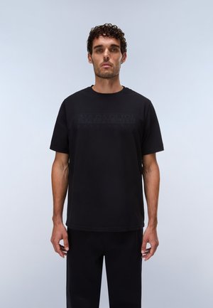 Mand med kort krøllet hår iført en ensfarvet sort t-shirt med korte ærmer og sorte bukser mod en lys grå baggrund.