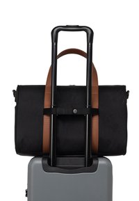 Bolsa de viaje de tela negra con asas de cuero marrón, asegurada a una maleta rígida gris mediante una correa negra. Presenta una textura suave.