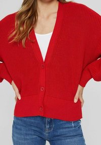 Rote Strickjacke mit V-ausschnitt, Knopfverschluss, übergroßen Ärmeln und geripptem Saum. Getragen über einem weißen Top und kombiniert mit blauen Jeans.