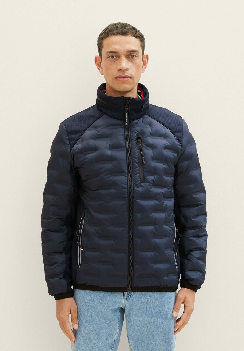 Giacca imbottita blu navy con colletto alto, tasca frontale con zip e polsini elasticizzati. Presenta un tessuto a contrasto sottile sulle spalle e ai lati.