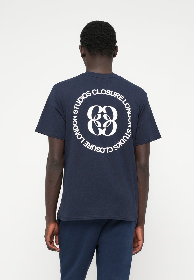 T-shirt de algodão azul-marinho com um grande logótipo circular branco com "STUDIOS CLOSURE LONDON" e design entrelaçado "83" nas costas.