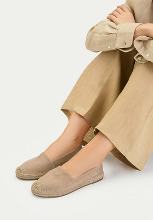 Beige Espadrilles mit geflochtener Jutesohle, getragen mit lockeren beigen Hosen; Person sitzt mit verschränkten Armen auf den Knien.