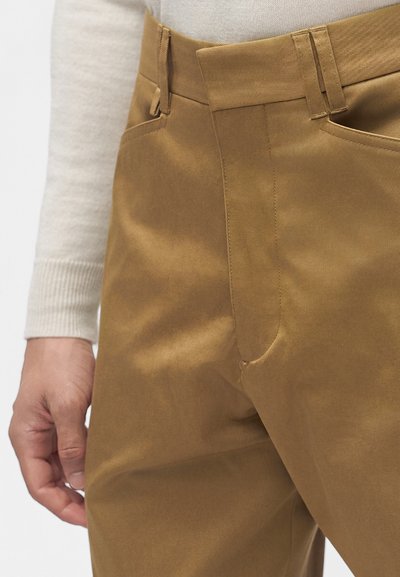 Gros plan d'une personne portant un pantalon marron avec passants de ceinture et poches latérales, associé à un haut à manches longues beige clair.