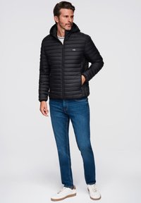 Chaqueta de plumas negra con cremallera frontal y capucha, textura acolchada horizontal, combinada con jeans de color azul claro y zapatillas blancas.