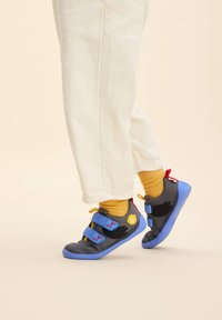 Zapatos de tela grises con suelas azules, con acentos de pájaros negros, un logo circular amarillo y correas de velcro dobles para un ajuste seguro.