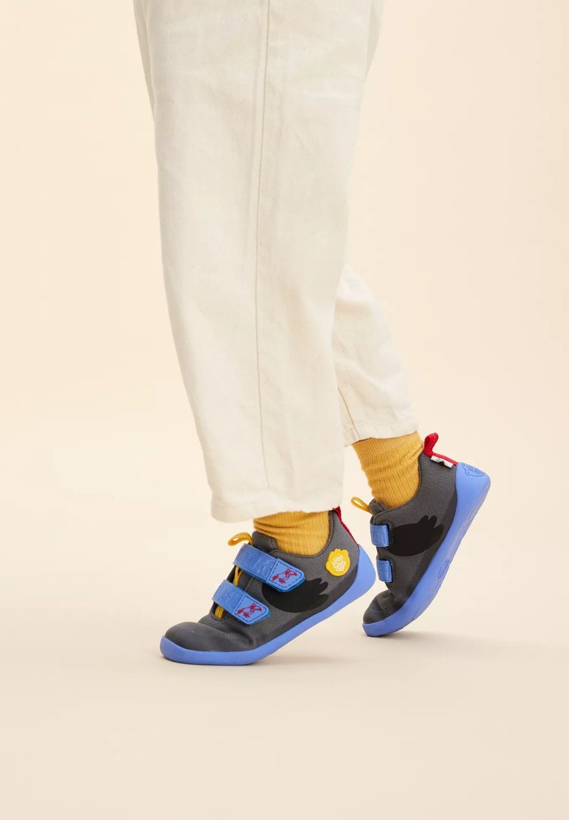 Zapatos de tela grises con suelas azules, con acentos de pájaros negros, un logo circular amarillo y correas de velcro dobles para un ajuste seguro.