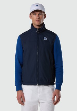 Homme portant une casquette blanche et un pantalon blanc, un gilet zippé bleu marine par-dessus une chemise à manches longues bleue, face à l'avant sur un fond uni.