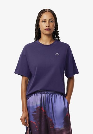 Camiseta de algodón morada con mangas cortas, cuello redondo y logo de Lacoste. Combinada con una falda estampada que presenta un diseño de paisaje en rosa y azul.