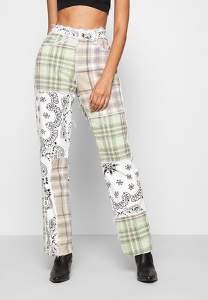 Vrouw die een broek met hoge taille draagt met patchwork van groene, beige ruiten en zwart-witte paisleyprints, gecombineerd met zwarte enkellaarsjes.