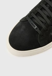 Sneaker in suede nera con punta arrotondata, superficie testurizzata e lacci neri piatti. Presenta una suola in gomma bianca e un logo sul lato.