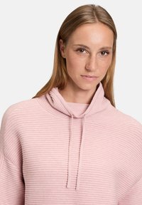 Jasnoróżowy sweter z kapturem, o prążkowanej strukturze i detalach w postaci sznurka. Posiada długie rękawy oraz luźny krój.