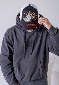 Grå huvtröja med framficka och snörning. Den svarta truckerkepsen har en färgglad broderad geisha-design och röd logotyp.