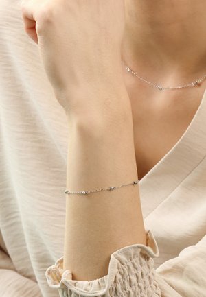 Silberne Kettenarmband mit kleinen runden Perlen, das ein zartes Design aufweist. Getragen an einem hellhäutigen Handgelenk, kombiniert mit einem sanften beigen Oberteil.