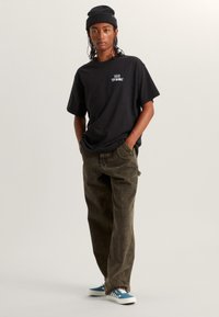 Camiseta de algodón negra con un logo en el pecho, combinada con pantalones holgados color verde oliva y zapatos a cuadros azules, usados con un gorro negro.
