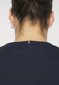 Námorně modré bavlněné tričko s kulatým výstřihem a viditelnou etiketou při krku, se stehy v červené, bílé a modré; hladká textura, ležérní design.