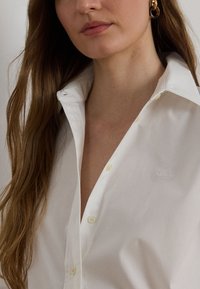 Žena s dlhými hnedými vlasmi nosí bielu košeľu na gombíky a zlaté kruhové náušnice, pričom sa zameriava na dolnú časť tváre a hornú časť trupu.