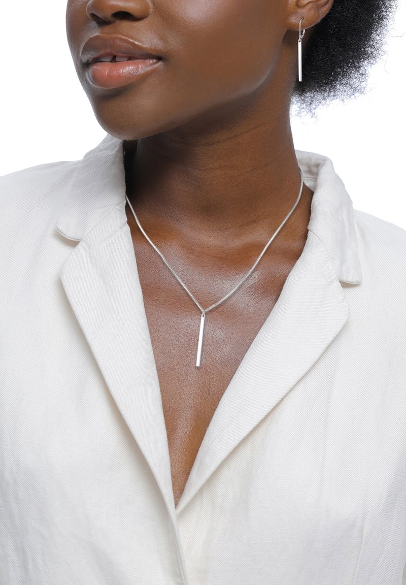 Sølvhalskæde med vedhæng i form af en lodret stang og matchende lange øreringe, båret af en person med mørk hud og naturligt hår, mod en hvid baggrund.