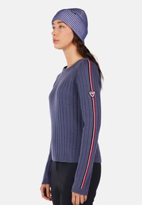 Fioletowy sweter w prążki z okrągłym dekoltem, z czerwonymi i białymi paskami wzdłuż rękawów oraz naszywką z logo na lewym ramieniu.