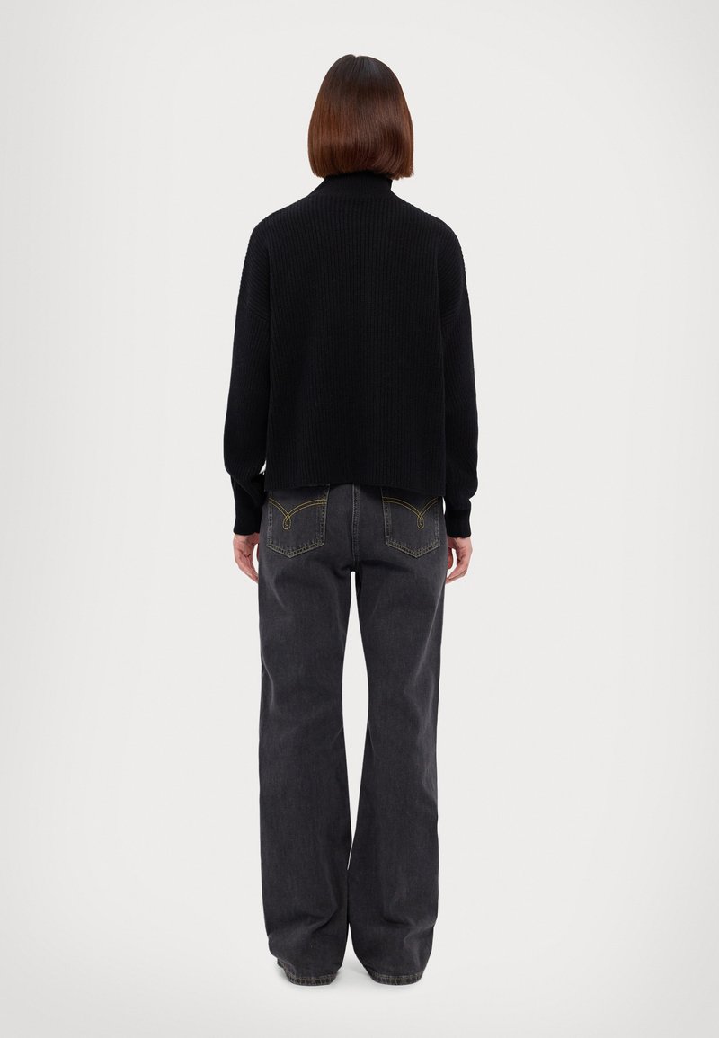 Schwarzer Strickpullover mit hohem Kragen, kombiniert mit lässigen, dunkelgrauen Jeans, die über Rückentaschen mit gestickten Details verfügen.