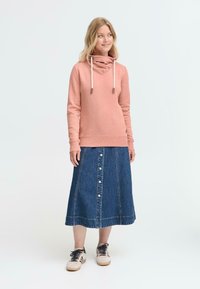 Felpa rosa con colletto a cowl e lacci, abbinata a una gonna di jeans lunga con bottoni frontali e taglio dritto. Calzature casual completano il look.