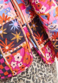 Kurtka pikowana z jaskrawym wzorem kwiatowym w odcieniach różu, pomarańczy i niebieskiego, z kontrastowym stębnowaniem i zaokrąglonym dołem.