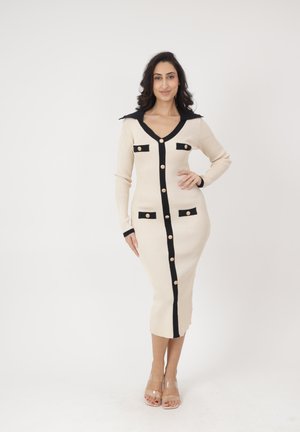 Frau posiert in einem figurbetonten, beigen Midikleid mit schwarzem Rand, goldenen Knöpfen, langen Ärmeln und Kragen, trägt transparente Riemchenabsätze vor weißem Hintergrund.