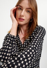 Zwarte en crèmekleurige blouse met patroon, voorzien van een V-halslijn, lange mouwen met knoopdetails en een lichte stof.