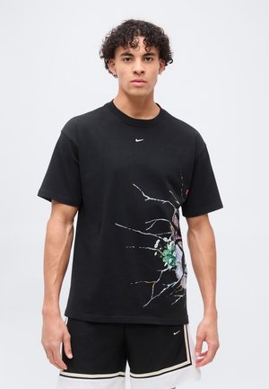 Jeune homme portant un t-shirt noir Nike avec un motif de verre brisé et une fleur sur le côté, assorti à un short noir et blanc, debout devant un fond uni.