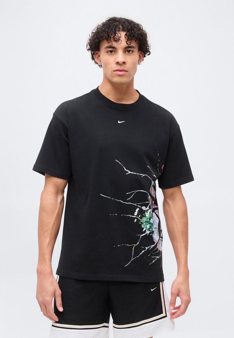 Jeune homme portant un t-shirt noir Nike avec un motif de verre brisé et une fleur sur le côté, assorti à un short noir et blanc, debout devant un fond uni.