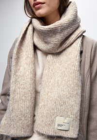 Sciarpa lavorata a maglia beige con texture a costine, morbida miscela di lana, con un'etichetta su un'estremità con la scritta "PARA STREET ONE" in testo nero.