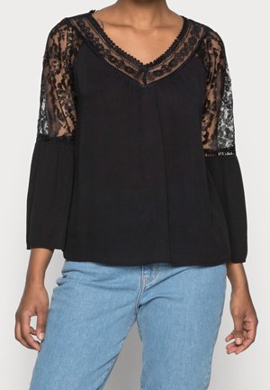 Blouse noire avec empiècement et manches en dentelle florale transparente, coupe ample, associée à un jean bleu clair.