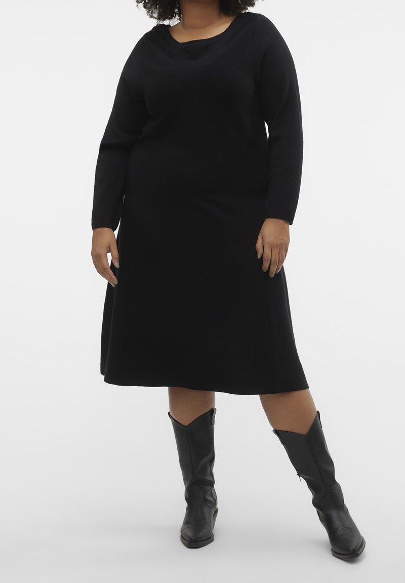 Femme portant une robe midi noire à manches longues associée à des bottes en cuir noires mi-mollet, posant devant un fond uni.