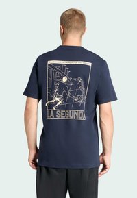 Marineblaues T-Shirt aus Baumwolle mit einer cremefarbenen Grafik auf der Rückseite, die eine Fußballsituation mit dem Text "LA SEGUNDA" zeigt.