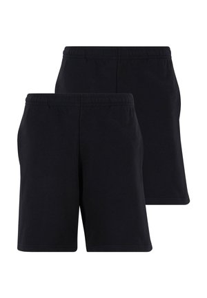 Personne portant un short noir, des chaussettes blanches montantes, des baskets blanches et noires, et un t-shirt blanc sur un fond clair uni.