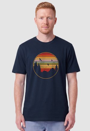 Mann mit kurzen roten Haaren, der ein dunkelblaues T-Shirt mit einem runden Sonnenuntergangsdesign trägt, auf dem Vögel und Bäume zu sehen sind.