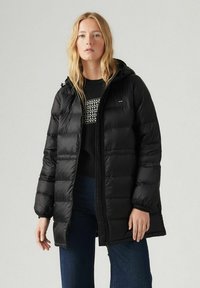 Chaqueta de plumas negra con cierre frontal y capucha, que presenta secciones acolchadas y puños elásticos. Combinada con un sudadera negra y unos vaqueros.