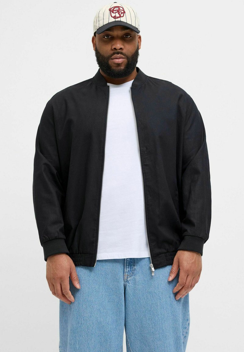 Schwarze leichte Jacke mit Reißverschlussfront, gerippten Bündchen und lockerer Passform, getragen über einem weißen T-Shirt und kombiniert mit hellblauen Jeans.