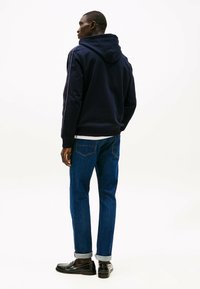 Marineblauwe hoodie met witte streepdetails, gecombineerd met blauwe denim jeans met opgerolde mouwen en zwarte loafers. Achteraanzicht weergegeven.