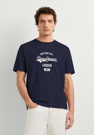 Ung mand iført en mørkeblå "Pepe Jeans London 1973" T-shirt, den ene hånd i lommen på hvide bukser, står mod en ensfarvet lys baggrund.