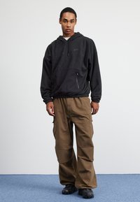 Črn flisast hup, ki ima kenguru žep, elastičen rob in vrvico; združen z ohlapnimi khaki cargo hlačami in črnimi supergami.