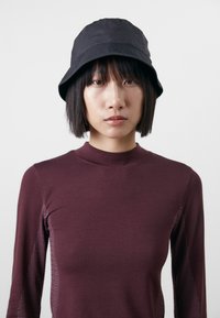 Svart bucket hat i lätt material med en rund topp och en bred, nedåtlutande skärm. Buren med en figurnära, långärmad vinröd topp.