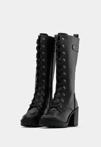 Pár černých kožených bot až ke kolenům s šněrováním vpředu, bočními zipy, robustními podpatky a profilovými podrážkami na jednoduchém bílém pozadí.