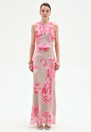 Donna che indossa un vestito senza maniche, lungo fino a terra, con grandi motivi floreali rosa su sfondo beige chiaro, orecchini e braccialetti dorati.