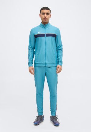 Mladý muž stojí, oblečený v svetlomodrej teplákovej súprave s čiernym pásom a bundou na zips, v kombinácii s čiernymi a fialovými športovými kopačkami.