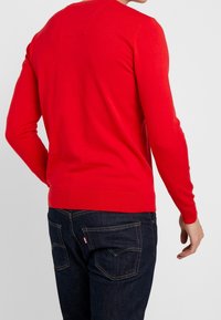 Maglione rosso a maniche lunghe con scollo rotondo e dettaglio patch sul retro. Indossato con jeans blu scuro, caratterizzati da cuciture a contrasto e dettagli sulle tasche.