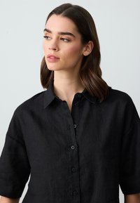 Blusa nera in lino con collo a camicia, maniche corte e chiusura anteriore con bottoni abbinati. Tessuto testurizzato con vestibilità rilassata.