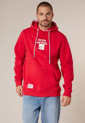 Homme portant un sweat à capuche rouge avec le texte « C'est la vraie chose. Coca-Cola », des cordons blancs et un jean bleu clair, sur un fond beige.